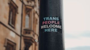 Trans people welcome here sticker on light pole. Letters are in trans flag colors.